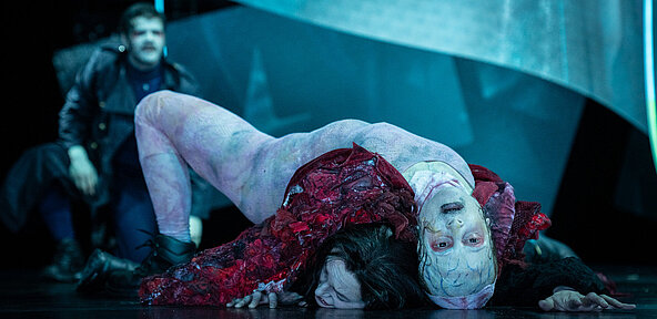 Frankensteins Geschöpf (Esther Berkel) liegt auf Viktor Frankenstein (Julia Friede) und drückt sie zu Boden. Es trägt einen rötlichen Hautanzug über Körper und Kopf und hat schwarze Augen. Im Hintergrund Robert Walton (Paul Enev).