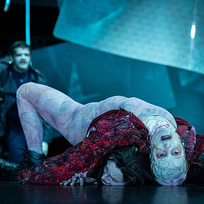 Frankensteins Geschöpf (Esther Berkel) liegt auf Viktor Frankenstein (Julia Friede) und drückt sie zu Boden. Es trägt einen rötlichen Hautanzug über Körper und Kopf und hat schwarze Augen. Im Hintergrund Robert Walton (Paul Enev).