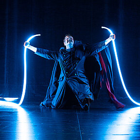 Robert Walton (Paul Enev) mit zwei hellen Lichtschläuchen in den Händen, die er triumphierend nach oben streckt. Dunkle Bühnenatmosphäre.