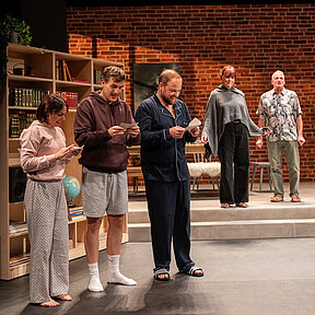 Luisa (Gesa Schierenstedt), Julius (Florian Pelzer) und Peer (Jan Wittje) nebeneinander in Schlafkleidung mit Schecks in den Händen. Im Hintergrund stehen Johanna (Maren Groth-Ische) und Winfried (Benno von Minden).