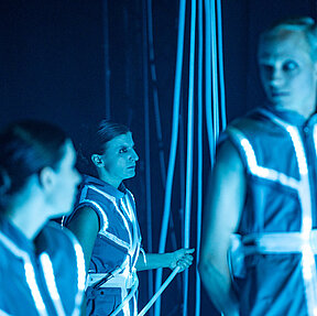 Vorne blicken Tamara Theisen und Tobias Schormann einander an. Dahinter: Katharina Shakina und hält ein langes weißes LED-Kabel, das von der Decke kommt