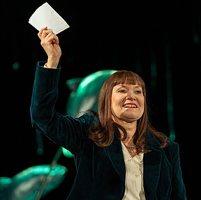 Martha Kiil (Caroline Nagel) hält mit der rechten Hand triumphierend ein Blatt Papier nach oben und lächelt. Sie trägt eine grüne Samtjacke, darunter eine weiße Bluse.