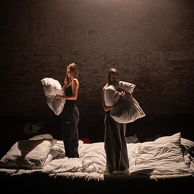 Zwei Darstellerinnen in Alltagskleidern stehen im Spot auf einer Matratze, mit dem Rücken zueinander. In ihren Händen weiße, große Kissen. 