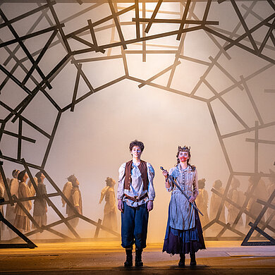 Hänsel (Dorothee Bienert) und Gretel (Stephanie Hershaw) stehen erleichtert vor einem Bogen. Gretel trägt eine Krone und hält ein Schwert, sie hat rote Farbe im Gesicht. Im Hintergrund der Kinderchor.