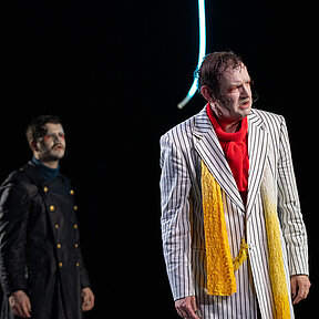 Alphonse Frankenstein (Klaas Schramm) blickt in die Ferne, er trägt ein rotes Oberteil und einen schwarz-weiß gestreiften Mantel mit gelbem Schal. Hinter ihm Elisabeth Lavenza (Esther Berkel) und Robert Walton (Paul Enev).