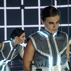 Katharina Shakina mit ernstem Blick Richtung Boden. Unscharf hinter ihr steht Tamara Theisen in nach vorne gelehnter Haltung. Graue Kostüme mit weiß leuchtenden LED-Streifen, hinter ihnen weißes Raster auf digitaler Wand. 