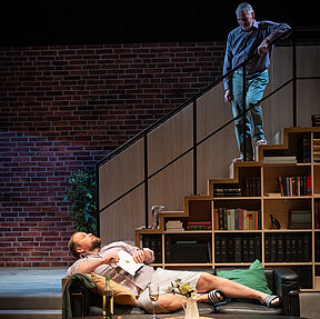 Peer (Jan Wittje) liegt auf dem Sofa und hält ein Tablet. Winfried (Benno von Minden) steht auf einer Treppe über ihm. Beide schauen sich an.