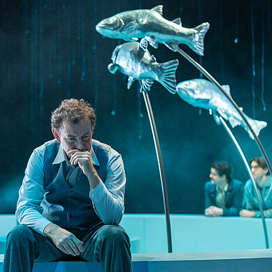 Tomas Stockmann (Klaas Schramm) sitzt nachdenklich auf dem Brunnenrand. Im Hintergrund mehrere Personen.