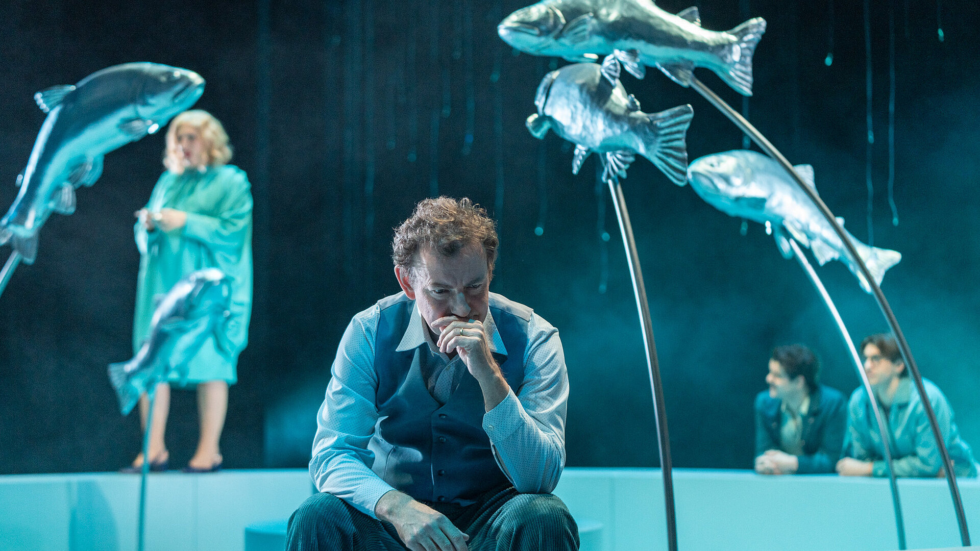 Tomas Stockmann (Klaas Schramm) sitzt nachdenklich auf dem Brunnenrand. Im Hintergrund mehrere Personen.