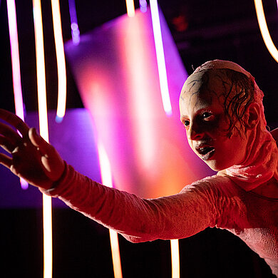 Frankensteins Geschöpf (Esther Berkel) in Nahaufnahme. Es streckt den rechten Arm nach vorne. Es trägt einen rötlichen Hautanzug über Körper und Kopf und hat schwarze Augen.