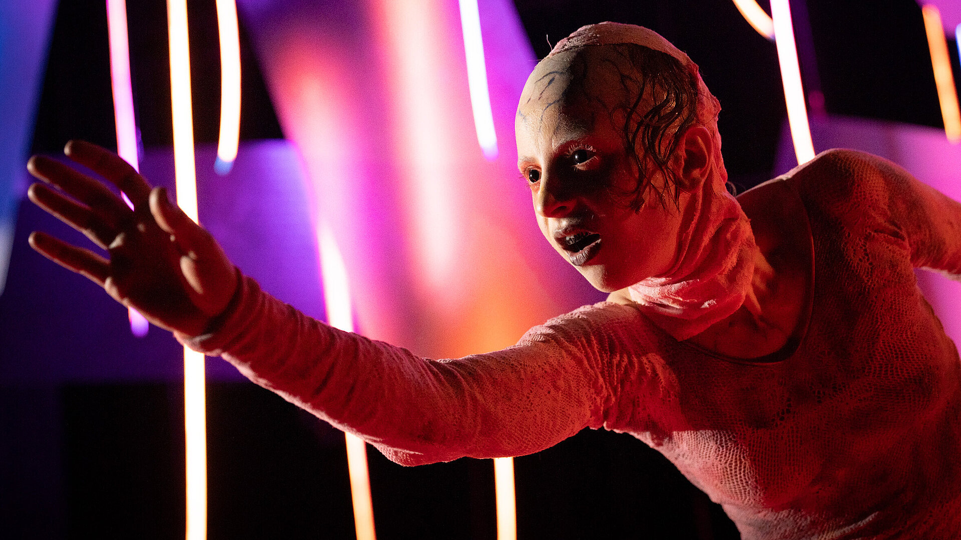 Frankensteins Geschöpf (Esther Berkel) in Nahaufnahme. Es streckt den rechten Arm nach vorne. Es trägt einen rötlichen Hautanzug über Körper und Kopf und hat schwarze Augen.