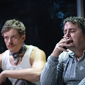 Fritz Hartmann (Gerrit Frers) raucht eine Zigarette. Im Hintergrund Carl Petersen (Florian Heise). Kühle, bläuliche Lichtstimmung.