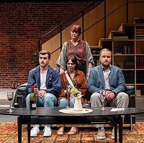 Julius (Florian Pelzer), Luisa (Gesa Schierenstedt) und Peer (Jan Wittje) sitzen nebeneinander auf einem Sofa. Auf dem Tisch vor ihnen eine Weinflasche, Weingläser und eine Blumenvase. Johanna (Maren Groth-Ische) steht hinter ihnen, Winfried (Benno von Minden), rechts neben ihnen.