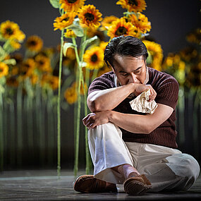 Alfredo Germont (Jason Kim), sommerlich gekleidet, sitzt mit überkreuzten Beinen und nachdenklichem, nach unten gerichtetem Blick, vor einem Sonnenblumenfeld, in seiner rechten Hand hält er einen zerknitterten Brief. 