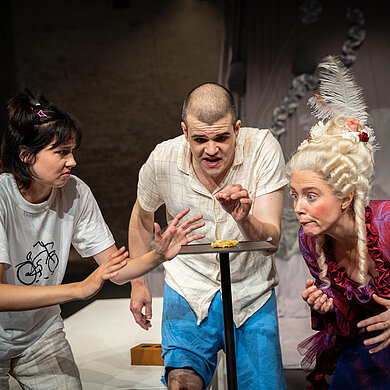 Die Geschwister Paula (Paulina Hobratschk) und Jonas (Paul Enev) sowie die Freifrau von Münchhausen (Cara-Maria Nagler) stehen im Halbkreis um den begehrten Keks herum und können die Augen nicht von ihm lassen.