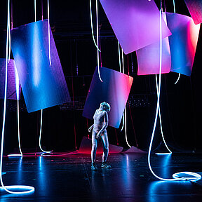 Frankensteins Geschöpf (Esther Berkel) blickt nach oben. Es trägt einen rötlichen Hautanzug. Um das Geschöpf hängende, halbtransparente Matten und hängende Lichtschläuche.