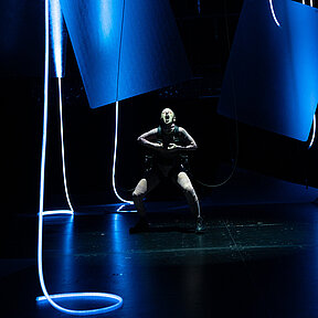 Frankensteins Geschöpf (Esther Berkel) in einer schreienden Geste, mit geöffnetem Mund. Es trägt einen rötlichen Hautanzug. Um das Geschöpf hängende, halbtransparente Matten und hängende Lichtschläuche.