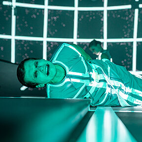 Konstantin Gries liegt mit schmerzverzerrtem Gesichtsausdruck und den Händen hinter dem Rücken auf dem Boden, türkises Licht. 