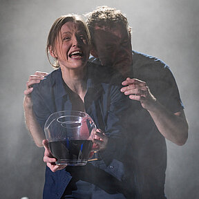 Zoe (Julia Friede) mit durchsichtiger Schüssel in den Händen, darin dunkle Flüssigkeit. Dennis (Klaas Schramm) hält Zoe von hinten an den Schultern.