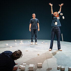 Fiffy (Franziska Werner) wirft weiße Holzklötzchen in die Luft. Um sie die anderen Schauspieler:innen.
