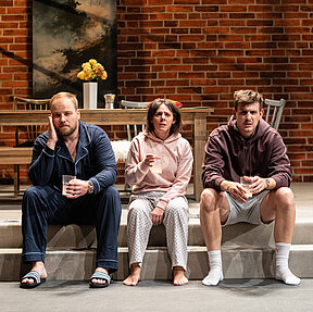 Peer (Jan Wittje), Luisa (Gesa Schierenstedt) und Julius (Florian Pelzer) sitzen verkatert auf Stufen. Sie tragen Schlafkleidung und halten Gläser mit Aspirin in der Hand.