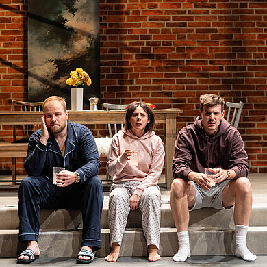 Peer (Jan Wittje), Luisa (Gesa Schierenstedt) und Julius (Florian Pelzer) sitzen verkatert auf Stufen. Sie tragen Schlafkleidung und halten Gläser mit Aspirin in der Hand.