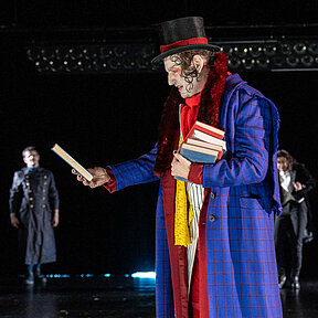 Professor Krempe (Klaas Schramm) in lila-rotem Mantel und schwarz-rotem Zylinder mit Bücherstapel. Er schaut sich ein Buch an. Im Hintergrund Robert Walton (Paul Enev) und Viktor Frankenstein (Julia Friede).