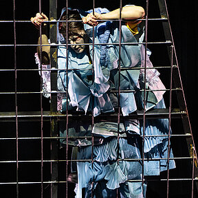 Justine Moritz (Katharina Shakina) hinter einem Gitter, an das sie sich klammert. Sie trägt ein blaues, plüschiges Kleid.