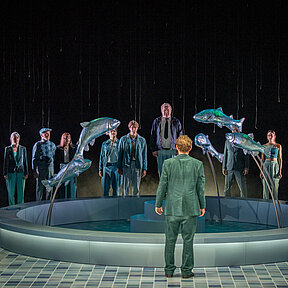 Tomas Stockmann (Klaas Schramm) mit dem Rücken zum Publikum. Das restliche Ensemble, in einer Reihe aufgestellt, mit Blick zu ihm.