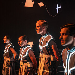 Spielende im Halbprofil, stehen in einer Reihe und blicken das Publikum spannungsvoll an. Das Licht ist warm und die LED-Streifen auf ihren Kostümen leuchten weiß. 