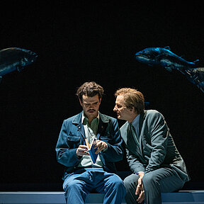 Peter Stockmann (Andreas Spaniol) bedrängt Redakteur Hovstad (Paul Enev). Sie sitzen, Hovstad blättert unkonzentriert in einem Buch. Düstere Lichtstimmung.