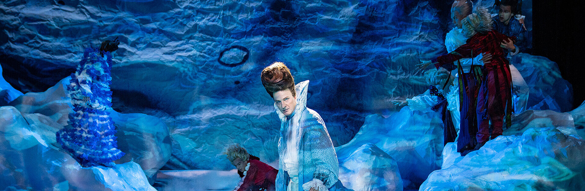 Prospero (Andreas Spaniol) steht vor der Felsenlandschaft. Rechts von ihm die Hofgesellschaft, die nach Ferdinand (Darios Vaysi) die Hände ausstreckt. Dieser liegt am Boden hinter Prospero.