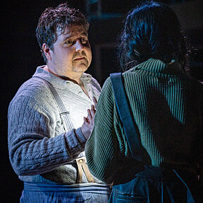 Fritz Hartmann (Gerrit Frers) in leicht blauem Kostüm mit Hosenträgern, blickt Nina Petersen (Paulina Hobratschk) an, die mit dem Rücken zur Kamera steht. Kühles Licht.