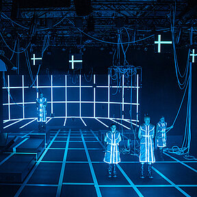 Vier Spielende stehen auf der Bühne, blau beleuchtet in der Totalen: Boden und digitale Wand hinten zeigen ein weißes Gitter. Durch den Raum hängen weiße Kabel. 