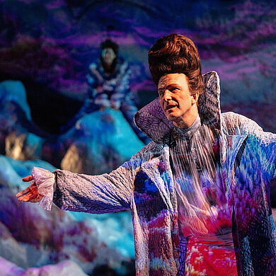 Prospero (Andreas Spaniol) hebt die Arme zu einer einladenen Geste. Er trägt seinen Zaubermantel.