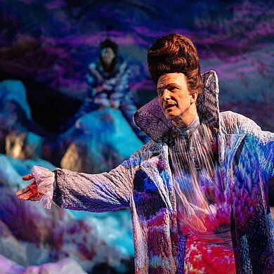 Prospero (Andreas Spaniol) hebt die Arme zu einer einladenen Geste. Er trägt seinen Zaubermantel.