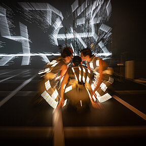 Ein unscharfes Bild: Vorne kommen die vier Spielenden hockend zusammen und die LED-Streifen auf ihren Kostümen leuchten warm und flirrend. 