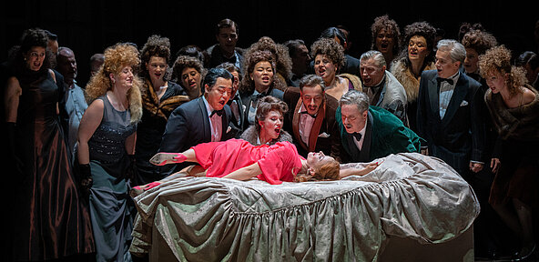 Violetta Valéry (Sarah Vautour) in einem roten weitfallenden langärmligen Kleid, auf einem weißen Bett. Sie wirkt unruhig und wird von zahlreichen Menschen angestarrt.