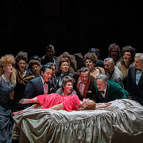 Violetta Valéry (Sarah Vautour) in einem roten weitfallenden langärmligen Kleid, auf einem weißen Bett. Sie wirkt unruhig und wird von zahlreichen Menschen angestarrt.