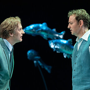 Peter Stockmann (Andreas Spaniol) steht Tomas Stockmann (Klaas Schramm) gegenüber, beide im Profil und mit Kostümen in Blautönen. 