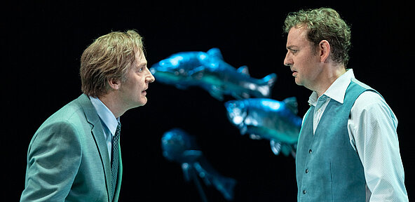 Peter Stockmann (Andreas Spaniol) steht Tomas Stockmann (Klaas Schramm) gegenüber, beide im Profil und mit Kostümen in Blautönen. 