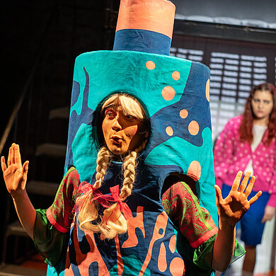 In Nahaufnahme ist eine stilisierte Litfaßsäule mit einer Shampoowerbung zu sehen. Aus einer Öffnung schaut das Gesicht des Spielers Finn Faust mit einer blonden Perücke und Zöpfen. Im Hintergrund ist unscharf Jeanne (Flora Reim) zu sehen.