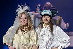 Zwei Darstellerinnen haben sich eingehakt und lächeln in die Kamera. Die eine hat eine Kappe mit Flügeln an der Seite auf. Die andere hat einen weißen Federpuschel auf dem Kopf.