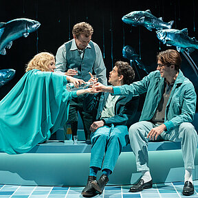 Katrine Stockmann (Meret Engelhardt), Tomas Stockmann (Klaas Schramm) und die Redakteure Hovstad (Paul Enev) und Billing (Jonah Winkler) schenken sich Schnaps ein. Hinter ihnen fünf große Fische an Metallstangen, die aus einem Brunnen herauszuspringen scheinen.