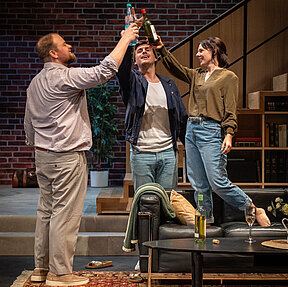 Peer (Jan Wittje), Julius (Florian Pelzer) und Luisa (Gesa Schierenstedt) stoßen an. Luisa steht dabei auf dem Sofa.