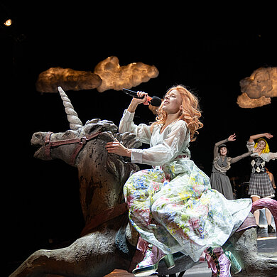 Jane Bennet (Esther Berkel) sitzt einem großen weißen blumigen Kleid und rosa Highheels auf einem weißen Einhorn und singt in ein Mikro. Über ihr leuchtet eine Wolke. Im Hintergrund winken weitere Spielerinnen.