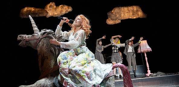 Jane Bennet (Esther Berkel) sitzt einem großen weißen blumigen Kleid und rosa Highheels auf einem weißen Einhorn und singt in ein Mikro. Über ihr leuchtet eine Wolke. Im Hintergrund winken weitere Spielerinnen.