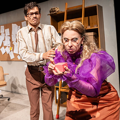 Lisa (Esther Berkel) steht vorne rechts und schaut mit aufgerissenen Augen auf ein kleines rosa Kästchen. Hinter steht Peter (Konstantin Gries) und hält sie an der Schulter. Er presst die Lippen zusammen. Im Hintergrund links steht Pico (Lukian Anhölcher) auf dem Aktenschrank.