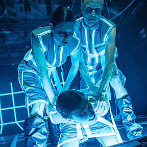 Tobias Schormann und Tamara Theisen drücken Konstantin Gries zu Boden. Ihre digitalen Kostüme leuchten blau.