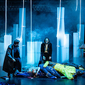 Viktor Frankenstein (Julia Friede) steht auf einem Berg voller Requisitenleichen. Robert Walton (Paul Enev) blickt ihn an. Im Hintergrund halbtransparente, aufrechtstehende Rollen, Nebel und leuchtende, hängende Schläuche.